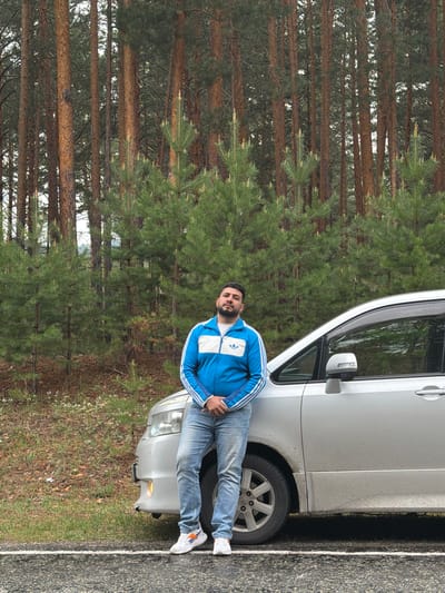 The image captures a man standing next to a silver van parked on the side of a road, with a dense pine forest visible in the background. The man is wearing a blue Adidas tracksuit and jeans. The forest is characterized by tall, straight pine trees. The location appears to be Сизая, Russia.