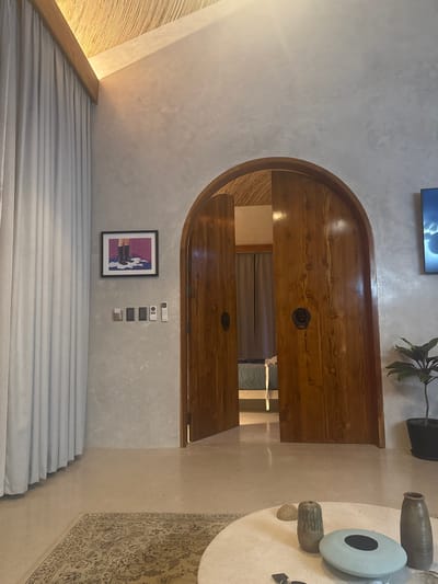 An interior view depicts a modern room, identified as being in El Nido, Philippines. The space features light grey textured walls and a vaulted ceiling, partially covered with a light brown, woven natural material. A recessed light strip runs along a wooden beam near the ceiling. On the left, floor-to-ceiling light-colored curtains obscure a window. A framed artwork, depicting legs in black boots standing in a white puddle against a purple background, hangs on the left wall. Below it, a series of wall-mounted controls includes three light switches and two remote controls.
Centrally positioned is an arched, double wooden door with a natural wood finish and dark lion-head door knockers. The right door is slightly ajar, revealing a glimpse of an adjoining room with a bed featuring a light blue comforter and a similar woven ceiling. To the right of the door, a dark-screen television displaying a light-colored, cloud-like image is mounted on the wall. Below the television, a potted plant with dark green leaves rests in a black pot on the floor.
The floor is a light-colored, smooth surface, possibly polished concrete. A patterned beige and brown rug with an intricate design is partially visible in the foreground. A round, light-colored coffee table occupies the lower right foreground, displaying various decorative items: a light brown cylindrical vase, a small irregular stone, a flat light blue oval object with a dark center, a taller dark brown ribbed vase, and a set of keys. No individuals are present in the scene.