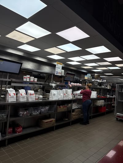 A fast-food restaurant setting shows a woman working behind the counter. She's packing food into bags, surrounded by multiple filled bags and organized supplies on metal shelves. POS systems and menu boards are visible above the counter. A tiled floor and a patterned ceiling provide context, hinting at the interior design.