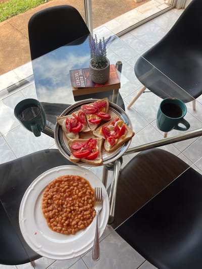 An overhead, static shot depicts an interior dining scene centered on a clear glass table, located in Bulawayo, Zimbabwe. Four modern chairs with dark seats are positioned around the table; one chair features visible wooden legs.

On the table, a white circular plate contains baked beans in tomato sauce, with a silver fork resting on its right. Adjacent to it, a striped plate (black and white rim) holds five slices of toast, each topped with a portion of egg and sliced red tomatoes. Two teal ceramic mugs, each containing a dark liquid, are placed on opposite sides of the table.

A small grey pot holding artificial purple lavender flowers is situated on two stacked books. The visible top book's spine reads "THE BURIED SECRET" by Katherine Graham.

The floor beneath the table is covered with light-toned tiles. Beyond a window, an outdoor concrete path and green grass are visible. Reflections of a cloudy sky are present on the glass tabletop, indicating natural overhead lighting. No individuals are depicted, and no actions or interactions are in progress.
