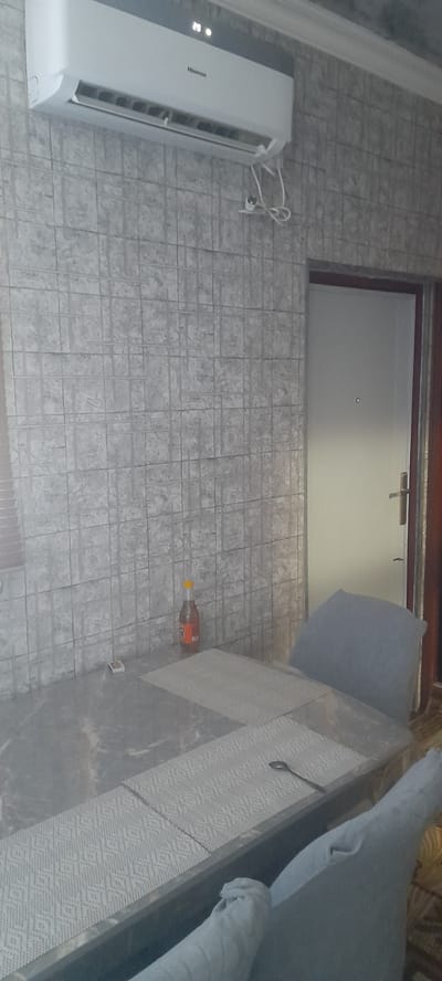 The image depicts an indoor scene, likely a dining area, within an apartment in Balkanabat, Turkmenistan. In the upper left portion of the frame, a white Hisense air conditioner is mounted on a wall covered with gray, grid-patterned wallpaper. A power cord is visible, connected to the air conditioner. Below the air conditioner, on a table with a marble-patterned surface, is a bottle of dark liquid with an orange cap, and a small rectangular object, possibly a box of matches. The table is set with placemats. To the right of the table, a portion of a door with a silver handle is visible. Two chairs, one mostly visible and the other partially obscured, are present. The chair on the right is covered with a light blue fabric. The overall lighting suggests a daytime interior setting.