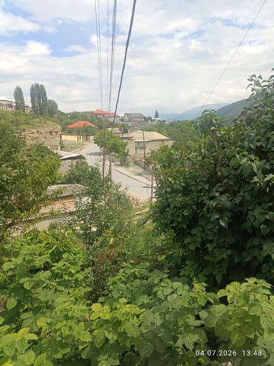 A scenic view of Rakhata, Russia, shows a street winding through a village nestled among rolling hills. Houses with red and beige roofs dot the landscape, surrounded by lush green trees and vegetation. The sky is partly cloudy, with a mix of blue sky and white clouds. Overhead, power lines crisscross the frame, indicating the presence of infrastructure. The foreground is dominated by dense green bushes, suggesting a natural, undeveloped environment. In the lower right corner, a timestamp "04.07.2026 13:48" is visible, indicating the date and time the photograph was taken. No people or animals are visible in the scene.