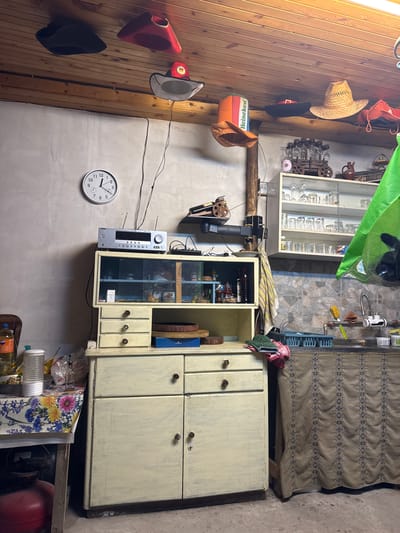 This appears to be a cluttered interior shot of a room, possibly in a home or storage area. The scene is a bit messy and has a rustic or lived-in feel.
**Key elements:**
* **Furniture:** There is a large, older cabinet with several doors. Next to it stands a smaller open cabinet with glass display cases.
* **Objects:** The room is filled with various objects including bottles, glasses, hats hanging on a wooden wall, tools, and electronic equipment like a sound system. A clock is on the wall, reading approximately 10:08.
* **Environment:** The walls are a mix of wood paneling and plastered surfaces. The floor appears to be a mix of concrete.
* **People and Actions:** The image does not show any people or ongoing actions.
Given that the room contains a "Heineken" themed item and a "FANTA" bottle, coupled with a slightly disorganized aesthetic, the setting is most likely a storage area or a room inside a home that has not been tidied.
While it is not confirmed where the photo was taken, there is a mention of Blajievo, Bulgaria, on the Heineken box.
