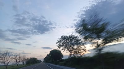 The image captures a scene from a moving vehicle along a road. The sky is filled with clouds, with a hint of sunset visible on the horizon. Trees line the roadside, some blurred due to the motion. A two-lane road stretches ahead, with a car's headlights visible in the distance. The location is unknown as the image does not provide the information that the location is in the city of Chernograd, Bulgaria.