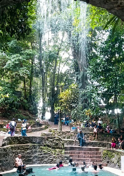The image depicts a natural bathing pool surrounded by lush greenery and large trees. Water cascades from an overhead structure, creating a gentle waterfall effect. Several people are present in and around the pool. In the pool, individuals are swimming and interacting with the water, with one person wearing a red swimsuit. On the stone steps leading into the pool, various people are seated or standing. Some are dressed in casual attire, while others wear hijabs. To the left of the pool, a person with a backpack stands on a stone pathway. Further back, more individuals are seated on stone ledges, observing the scene. The environment is tropical, with dense foliage and vines hanging from the trees. The location is identified as a natural bath in Penungkiren, Indonesia, suggesting it's a recreational or tourist site.