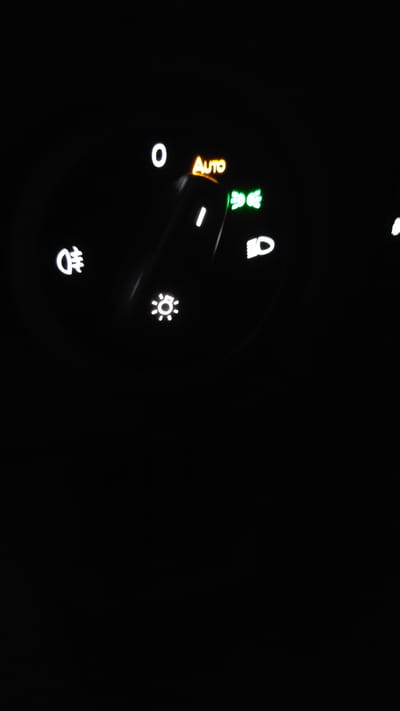 The image displays a close-up of a car's illuminated light control dial, set against a completely black background, indicating a dark environment or nighttime conditions in Westerlo, Belgium. The central control knob is partially visible, with a prominent white indicator line pointing to the "AUTO" setting. The word "AUTO" is illuminated in orange, signifying that the automatic light mode is selected. Next to "AUTO," a green symbol depicting headlights with light rays is also illuminated, indicating that the automatic headlight system is active.

Around the dial, several other lighting functions are represented by white illuminated symbols:
- A "0" indicates the lights-off position.
- A symbol for low beam headlights (a headlight icon with downward-sloping light rays).
- A symbol for front fog lights (a headlight icon with downward rays crossed by a wavy line).
- A symbol resembling a sun or starburst with radiating lines, typically representing parking lights or sidelights.
- A symbol depicting a headlight with upward-sloping rays and a squiggly line, which commonly signifies rear fog lights.

The bright and crisp illumination of all symbols and text against the dark backdrop ensures clear visibility and readability of the car's lighting controls.