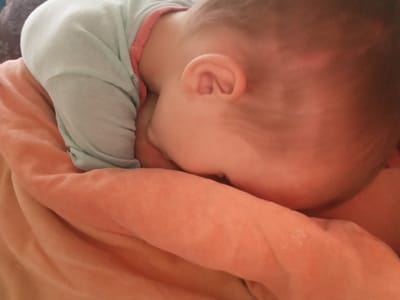 A close-up, warm-toned photograph depicts a baby or young child sleeping or resting peacefully. The child is positioned face down, snuggled into a soft, textured, orange-peach colored blanket or fabric. Only the back of the child's head, the right ear, and a portion of their upper back and arm are visible.

The child is wearing a light blue long-sleeved garment, which has a contrasting pink or reddish trim around the neckline or collar. The ear is clearly visible, showing its delicate convolutions. The child's skin appears smooth and healthy. The lighting is soft, creating a gentle and intimate atmosphere, suggesting an indoor setting, likely a quiet space like a nursery or bedroom in Teykovo, Russia. The focus is slightly soft throughout the image, contributing to the tranquil mood. There is no visible text in the image.