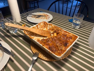 The image shows a partially eaten dish of lasagna in a white rectangular baking dish on a green and white striped tablecloth. There is a wooden spatula lying on the table next to the dish, and a small woven placemat under the dish. There is also a clear glass of water and a partially eaten white plate on the table. The location is in Blåvand, Denmark, at the coordinates 55.556, 8.138.