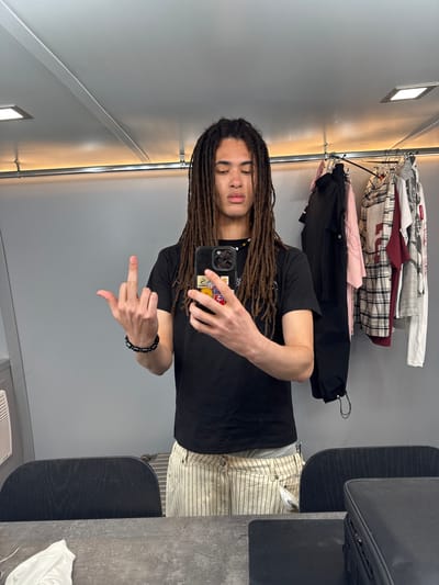 A medium shot, taken at an angle, shows a man with dreadlocks in an interior space. The man is wearing a black t-shirt and striped pants. He is holding a phone, taking a selfie, and extending his middle finger. A clothing rack with various shirts and jackets is visible in the background. The interior is well-lit with gray walls and metallic accents. There is no indication of this image being taken in San Bartolomé, Spain.