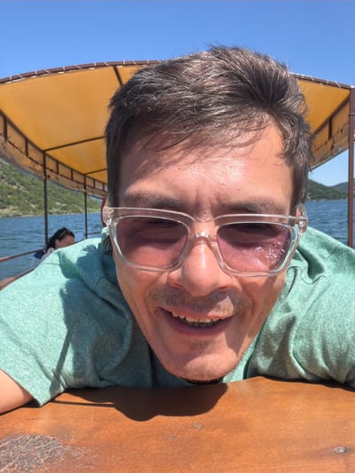 The image presents a close-up, selfie-style photograph of an adult male situated on a boat in Kruševica, Montenegro, under a clear, bright blue sky.
The primary subject, an adult male, occupies the lower two-thirds of the frame, looking directly into the camera. He has short, dark brown hair, with some grey strands visible, and a light mustache and stubble on his chin and jawline. His skin is tanned, exhibiting visible pores and a slight sheen on his forehead. He is wearing a light green, textured short-sleeved shirt. Clear-framed sunglasses with dark, possibly purple-tinted lenses, bearing the "GUESS" brand on the right temple, cover his eyes. He is smiling broadly, revealing his lower teeth. His forearms and upper torso are resting on a weathered wooden surface in the immediate foreground.
In the mid-ground, behind the main subject, another individual, partially obscured and appearing female, is visible to the left, facing away from the camera. This person has dark hair and wears sunglasses perched on their head, along with a white top. Above the subjects, a yellow/orange canvas canopy, supported by a dark brown frame, forms part of the boat's overhead structure.
The background features a large body of water with ripples, stretching towards distant green, tree-covered hills or mountains that rise from the water's edge. The sky above is entirely clear and bright blue. Strong sunlight from an elevated position casts distinct shadows under the canopy and on the wooden surface in the foreground, as well as subtle shadows on the main subject's face.
