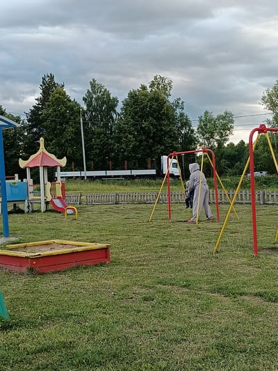 The image shows an outdoor playground with two people on a swing set. One person, wearing a grey hooded tracksuit, is pushing a child on a swing. The playground equipment includes the swing set, a sandbox with a red and yellow border, and a small play structure with a slide. In the background, there are trees, a wooden fence, and a white truck. The sky is cloudy, suggesting it might be late afternoon or early evening. The scene appears to be in a rural or suburban area, possibly a park or a residential backyard, given the presence of trees and a fence. There is no visible text in the image.