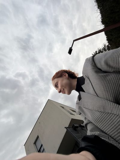 The image depicts an outdoor scene in Eschau, France, under an overcast sky characterized by widespread light grey clouds. A female subject with reddish-brown hair, wearing a black turtleneck and a grey and white houndstooth patterned blazer, occupies the right foreground and mid-ground. She is positioned looking downwards and to the left, with her lips slightly upturned at the corners. Her arm is partially visible along the bottom edge of the frame. To the left of the subject, a section of a modern building with light-colored, possibly beige or off-white, walls is visible. This building features dark-framed windows and a dark balcony structure. In the upper right quadrant, a dark pole extends diagonally upwards from behind dense green foliage, terminating in a downward-bent section from which a small, dark, rectangular electronic device is suspended. The photograph is taken from a low-angle perspective, looking upwards towards the sky and background elements.