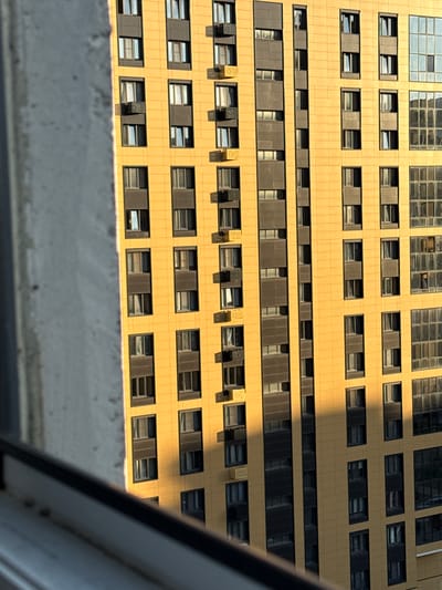 The image shows a close-up, angled view of a modern, yellow, multi-story apartment building, viewed from a window. The building features a repetitive pattern of windows and dark-colored recessed panels. The sun is casting a shadow across the building from the right side. In the foreground, a portion of a gray, textured structure, likely part of the adjacent building or a window frame, is visible on the left, partially obscuring the view of the yellow building. The image is taken outdoors, and the overall impression is one of urban architecture with a strong emphasis on geometric design and the play of light and shadow. There are no people or specific actions visible in the frame. The image appears to have been taken in Podkletnoe, Russia, judging by the prompt's request.