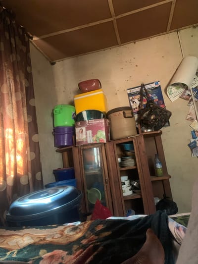 The image depicts a cluttered interior room. In the foreground, a large black basin is placed on a patterned blanket or bedspread. To the left, a brown and white polka dot curtain is visible. A wooden shelf unit stands in the center, filled with various items including stacked plates, pots, and bottles. Several plastic containers of different colors are stacked high next to the shelf unit, including green, purple, yellow, and red. A large white cylindrical object, possibly a speaker or a container, is leaning against the wall on the right. Several papers and posters are affixed to the wall behind the shelf unit. The walls appear aged and stained, and the ceiling is constructed with wooden planks. The scene suggests a domestic setting, possibly in a residential area of Gwolong, Nigeria, characterized by the presence of everyday household items and storage solutions. There are no people clearly visible in the image.