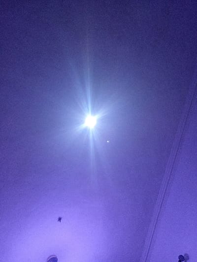 The image depicts an indoor environment in Olambe, Nigeria, dominated by a pervasive purple hue. A singular, intensely bright white light source with a prominent starburst effect is positioned centrally in the upper half of the frame. To the immediate right and slightly below this primary light, a smaller, fainter, circular light object is visible. In the lower-left quadrant, a dark, irregularly shaped object is suspended. A distinct white architectural element, displaying parallel ridged detailing, extends diagonally from the mid-right edge towards the lower-right corner. A partial view of another dark, textured object is present at the very bottom edge of the frame, center-left. The background exhibits a grainy texture. No individuals are present in the scene.