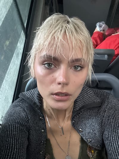 A close-up selfie taken indoors, likely on public transportation, shows a young woman with short, wet, blonde hair and light green eyes. She is wearing a dark grey knitted zip-up jacket over a dark green garment and several silver necklaces. Her face is turned towards the camera, and her expression is neutral. The background is out of focus, but appears to include a window with water droplets on it, suggesting it might be raining. To the right of the woman, another person wearing a red jacket and holding a clear plastic bag is partially visible. The lighting is somewhat dim. The image does not provide enough information to determine the specific location within Changning District, China.