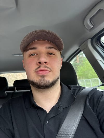 A man with a beard and a brown cap is sitting in a car. He is wearing a black shirt and a seatbelt. The interior of the car is gray. The exterior view through the car window shows trees and some kind of white railing. The photo was taken in Byala Reka, Bulgaria.