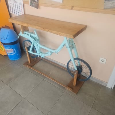 A light blue bicycle frame has been repurposed as a table base. A wooden plank rests on top of the frame, serving as the tabletop. The table is positioned indoors, likely in a commercial or communal space. To the left of the table, a blue plastic trash can is visible, and further left, a portion of an orange object. On the wall behind the table, a calendar with "JULIO 2026" prominently displayed hangs, along with other papers. A white electrical outlet with a smiley face sticker is on the wall to the right. The floor is tiled with large, grey square tiles. The overall impression is that this is a functional yet decorative piece of furniture, possibly located in a public area of Barrio Estación, Spain.