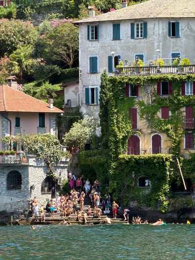 The image depicts a lively scene at a lakeside public area in Nesso, Italy. In the foreground, the rippling green water of Lake Como is visible, with several individuals swimming and relaxing. A person on an orange inflatable float is positioned in the mid-ground of the water.
The focal point of the image is a stone staircase leading from the water up to buildings adorned with lush green ivy. A large crowd of people, dressed in swimwear, are seated and standing on these steps, socializing and enjoying the sunny weather. Some are interacting with each other, while others are looking out at the lake.
Behind the steps, two buildings are prominently featured. The building on the left has a reddish-brown tiled roof and light-colored walls, with arched windows. The larger building on the right is a multi-story structure covered in dense green ivy. It has multiple windows with dark shutters and a balcony overlooking the lake, where a few people are visible. The architectural style suggests historic European lakeside residences. The background consists of a steep, verdant hillside covered in trees and flowering bushes, characteristic of the Italian lake region. The overall atmosphere is one of a popular summer gathering spot.