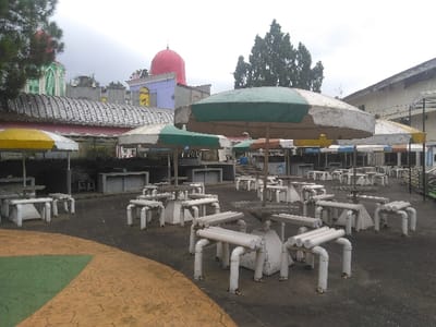 The image displays an unoccupied outdoor dining or recreational area located in the city of Ede, Netherlands, under an overcast sky. The foreground shows a dark paved surface with a section of patterned brown concrete and an adjacent green, smooth concrete area. The primary subjects are numerous white, weathered tables and benches. The tables are square or octagonal, accompanied by benches constructed from white-painted tubular elements, exhibiting paint chipping and dirt accumulation. Overhead, multiple large parasols, faded in segments of green, white, yellow, and brown, provide shade. In the background, a series of buildings is visible. To the left, a structure with a green and white facade, arched windows, and a small dome is present, next to another building featuring a prominent red dome atop a light purple section with yellow architectural accents. A long, curved roof extends across the mid-ground. To the right, a larger, light-colored building with a dark, sloped roof is partially visible. A cluster of dark green coniferous trees is situated in the mid-right background. No individuals are present in the scene, and no actions or interactions are depicted.