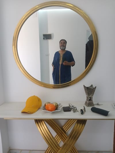 The image shows a man standing in front of a large, round, gold-framed mirror in what appears to be a modern home interior. The man is wearing a dark blue robe with gold trim and is holding a smartphone. His reflection is visible in the mirror, which also reflects a white wall with some electrical outlets and a framed picture.
On a console table below the mirror, there are several items:
- A yellow baseball cap with "NIKE SPORTS" written on it.
- A small, decorative silver tray containing two gold-colored spherical ornaments and an orange, egg-shaped object.
- A pair of eyeglasses.
- A dark brown wallet.
- A dark green cylindrical object, possibly a pen holder or a case.
- An ornate, silver incense burner.
- A set of keys.
The console table itself has a white marble top with gold trim, and its base is made of intersecting gold-colored metal beams, forming an "X" shape. The flooring is not clearly visible, but appears to be tiled. The overall impression is one of a well-decorated, contemporary living space. The location is specified as Hun, Libya.