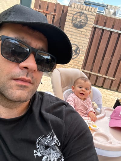 The photograph depicts a man and an infant in an outdoor setting. The man, wearing a black baseball cap and sunglasses, is positioned in the foreground, likely holding the camera for a selfie. He is wearing a black t-shirt with a white dragon design. Behind him, an infant is seated in a high chair with a pink hat on top of it. The infant wears a pink long-sleeved shirt. A toy rests on the high chair's tray. A wooden fence and the exterior of a brick building are visible in the background. Decorative metal pieces adorn the brick wall. The reflection in the man's sunglasses reveals the presence of buildings and sky, indicating an urban location. Based on the available information, this image was taken in Veghel, Netherlands. The image appears to be a casual family snapshot taken outdoors.