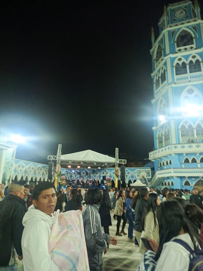 The image depicts a nighttime scene in El Cisne, Ecuador, featuring a crowd of people gathered in front of a brightly illuminated, multi-story blue and white church tower. A stage is visible in the background, partially covered by a white canopy, with a band performing. The foreground shows a group of people, including a man in a white hoodie holding a pastel-colored blanket. The crowd seems to be diverse in age and attire. The lighting conditions suggest artificial illumination from the stage and the church tower. The architectural style of the church tower is ornate, featuring arched windows and a clock at the top.