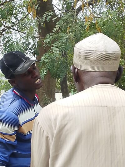 In Daurawa, Nigeria, two adult males are depicted outdoors. The individual on the left, facing right and slightly away from the camera, is dark-skinned, wears a black baseball cap, and a long-sleeved polo shirt with horizontal stripes in blue, light blue, white, and beige. His mouth is open, revealing upper teeth, indicating speech or expression directed towards the second individual. The individual on the right, observed from the back of the head and upper torso, is also dark-skinned, wearing a light-colored, patterned woven skullcap (kufi) and a light beige or off-white collared shirt with faint vertical stripes. His ears are visible. Both individuals appear engaged in a face-to-face interaction. The background features dense green foliage from trees, with a visible tree trunk and several elongated, light brown seed pods hanging from branches, suggesting an outdoor, possibly park-like or roadside, setting under daylight conditions.