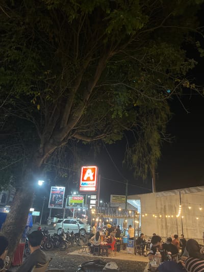 The image depicts an outdoor gathering at night in Tambonbaroh, Indonesia, likely a food court or a street-side eatery. A large tree with dense foliage dominates the left side of the frame, its branches casting shadows. In the background, a brightly lit "Alfamidi" sign is visible, indicating a nearby convenience store. Several other signs, including one for "INDO HOUSEWARE," are also present.
The scene is populated by numerous people seated at tables and chairs, engaged in various activities such as eating, drinking, and socializing. The atmosphere is illuminated by strings of warm, yellow lights that create a festive ambiance. Motorcycles are parked in rows along the side, suggesting a mode of transportation for some patrons. A silver SUV is parked further back, near the Alfamidi store. The overall environment appears to be a bustling, informal urban setting at night.