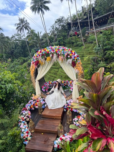 A single adult female is depicted seated within a large, elaborate floral archway on a tiered wooden platform in Tegal Suci, Indonesia. She wears a long, light-colored dress, a beige wide-brimmed hat, and sunglasses, looking upwards and slightly to her left. The archway, constructed of natural fibers, is heavily adorned with artificial flowers of varied bright colors, including red, blue, pink, yellow, and orange, and features cream-colored drapes tied back. Several golden-hued lanterns are placed along the wooden steps, amidst additional floral decorations. The background reveals a tropical landscape with dense green vegetation, terraced rice paddies, numerous tall palm trees, and a multi-level building with dark roofing integrated into a distant hillside. Utility lines are discernible in the sky. Foreground elements include large, reddish-green-leafed plants on the right and diverse green foliage on the left.