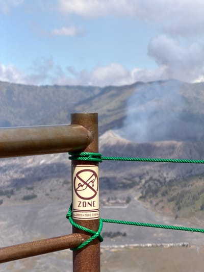The image depicts a close-up view of a metal fence post with a sign attached, in the vicinity of Mount Bromo, Cemorolawang, Indonesia. The sign reads "ZONE" and has a red circle with a diagonal line across it, superimposed on an image of a person wearing hiking boots, indicating a restricted area. Below "ZONE" is smaller text that appears to say "ADVENTURE TRAIL". A green rope is tied around the fence post, securing a section of the fence. In the background, a hazy, mountainous landscape is visible, with a plume of smoke rising from what appears to be a volcanic crater. The sky is partly cloudy, with blue sky visible between the clouds. The scene suggests a volcanic observation point or a trail leading towards a volcanic area. There are no people visible in the immediate foreground.
