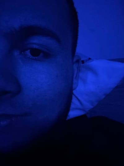 The scene depicts a close-up view of the left side of a human face and upper torso, illuminated predominantly by a strong blue light, occurring indoors in the city of Nadarevo, Bulgaria. The individual appears to be a male with short-cropped hair. The left eye is open, pupil visible, directed towards the viewer. A dark, arched eyebrow is prominent above the eye. Portions of the nose, left cheek, and upper lip are discernable. The individual's left ear is partially visible along the right edge of their head. To the right of the individual, a crumpled white fabric, resembling a pillow or bedding, rests against a similarly blue-lit wall. A dark, indeterminate fabric, possibly bed covers, is visible in the lower right foreground. The subject appears to be in a reclining position. No other subjects, objects, or specific actions are depicted beyond the subject's pose and environment.