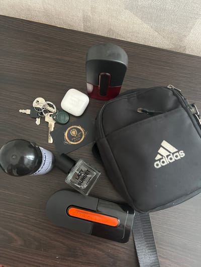 The image displays a collection of personal items laid out on a dark wooden surface, likely a desk or table. The items are arranged in a somewhat scattered manner. In the foreground, there is a black cylindrical object with a white label, a glass perfume bottle with a black cap and the label "LAZY SUNSET," and a black electronic device with an orange accent. Behind these, a set of keys with various keychains is visible, alongside a white wireless earbud charging case. To the right, a black crossbody bag with the Adidas logo is prominent. In the upper portion of the image, a black and red object, possibly a stamp or a dispenser, is positioned. The background appears to be a textured wall. The scene is static, with no discernible actions or interactions taking place. The lighting suggests an indoor environment. The location is not visible in the image, and there is no information to suggest it is in Sobishchytsi, Ukraine.