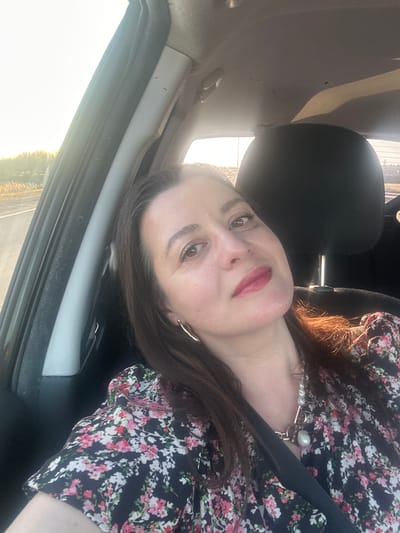 A woman with brown hair and light skin is sitting in the driver's seat of a car. She wears a floral-patterned blouse and a pearl necklace. The car interior is visible, including the black headrest and the car's frame. Outside the window is a bright, hazy scene suggesting either sunrise or sunset. The location is specified as the city of Piņķi, Latvia, but this is not visually verifiable from the image.