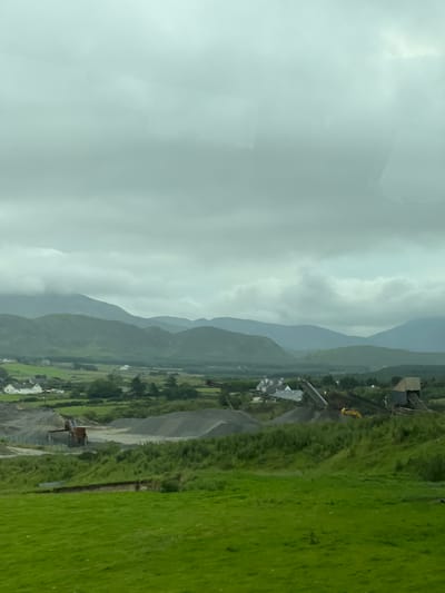 This media file depicts a vast, mountainous landscape under a cloudy sky, suggesting a cool or temperate climate. The scene is oriented vertically, with the mountains stretching towards the top of the frame. In the foreground, a vibrant green grassy field occupies the right side, contrasting with the muted tones of the distant mountains and sky.
Nestled within the valley between the mountains and the field is a small collection of buildings, hinting at a rural or semi-rural settlement. A construction site is visible, with heavy machinery, including an excavator, present. Piles of gravel or dirt are also evident, indicating ongoing work or development. The presence of the construction site suggests human activity and industry within this otherwise natural setting.
The weather appears overcast, with diffuse light indicating it might be daytime, possibly late afternoon or early morning given the soft lighting. There are no people clearly visible, but the constructed elements and machinery imply their presence. The rolling hills and green fields are characteristic of a place like Fanmore, Ireland, known for its scenic beauty and agricultural land. The overall mood conveyed is one of natural grandeur intersected by human endeavors, under a potentially moody, Northern European sky. No visible text is present in the image.