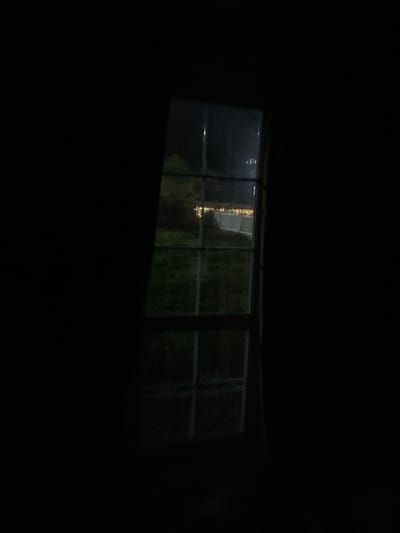 The image shows a window at night. Outside the window, there is a dim cityscape, likely in the Хлябово area of Russia, based on the prompt. A string of lights can be seen through the window. The inside of the room is dark.