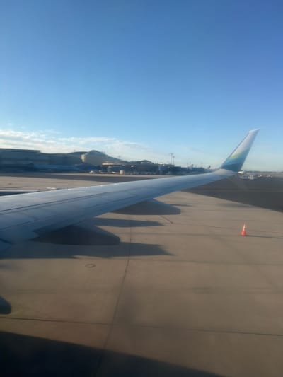 An elevated view from an airplane window shows an airport tarmac under a clear blue sky. The prominent subject is the airplane wing, extending from the bottom left corner and stretching across the lower half of the frame. The wing is metallic and features a distinctive teal and green stripe design on its vertical stabilizer, which is visible in the upper right portion of the image.
In the background, various airport structures are visible, including terminals, hangars, and other parked aircraft. Some of these structures appear to be modern buildings with large glass facades. The tarmac itself is paved with concrete and marked with yellow and red lines. An orange traffic cone is situated on the tarmac in the middle right of the image. The overall scene suggests the airplane is either preparing for departure or has recently arrived at the airport. The location is identified as Newark Liberty International Airport (EWR) in Newark, New Jersey, United States of America.