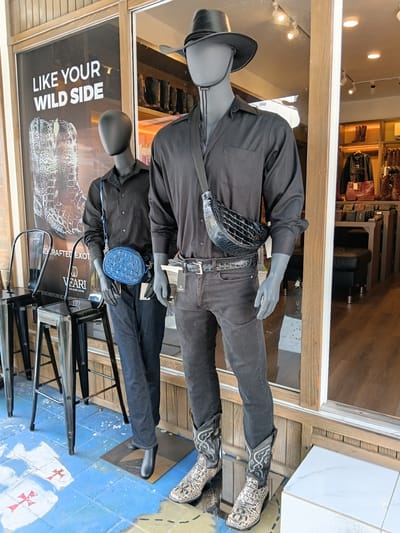 This is a vertical, eye-level, full-shot photograph of two mannequins dressed in Western-style attire, displayed in what appears to be a store window. The mannequins are positioned one above the other, both facing forward. The upper mannequin wears a black button-down shirt, black jeans, a black cowboy hat, and patterned cowboy boots. A black, textured fanny pack is worn across its chest. The lower mannequin is dressed similarly, with a black shirt and dark jeans, and is accessorized with a small, blue, quilted handbag with a strap.
The background consists of a store interior, featuring shelves stocked with various leather goods, including handbags and shoes. The floor in front of the mannequins is tiled in blue and white, with a red cross design visible on some tiles. A partial view of a poster with text is visible behind the lower mannequin. The overall scene suggests a retail display, possibly in a boutique specializing in leather fashion. The lighting appears to be artificial, typical of an indoor setting. No specific time of day or weather conditions can be determined from the image.
Visible text on the poster behind the lower mannequin includes "LIKE YOUR WILD SIDE" in large white capital letters, followed by "CRAFTED EXOT..." and other text in smaller fonts, including "WEAR I" and "REPUBLIC LEATHER."