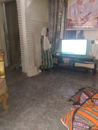 An elderly woman wearing a hijab and a striped dress stands in a living room next to a fluted pillar. A TV screen with a bright display sits atop a dark wooden stand against a window. A poster hangs above the TV. Curtains frame the window. The floor is covered in patterned tiles. A colorful patterned rug lies near the bottom right corner. Part of a gold-colored chair is visible on the left. A power outlet with a charger is on the wall next to the TV.