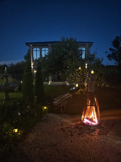 The image depicts a backyard scene at dusk in Надежда Снт, Russia. A modern, two-story building with large arched windows is visible in the background. The foreground features a lit fire pit with burning logs, surrounded by gravel. To the left of the fire pit, a wheelbarrow rests on the ground. The yard is illuminated by small, yellow solar lights scattered throughout the grass and a string of fairy lights draped around a tree, which also has decorative yellow lanterns hanging from its branches. A dark blue, clear sky is visible above. The overall impression is of a tranquil, evening outdoor gathering space.