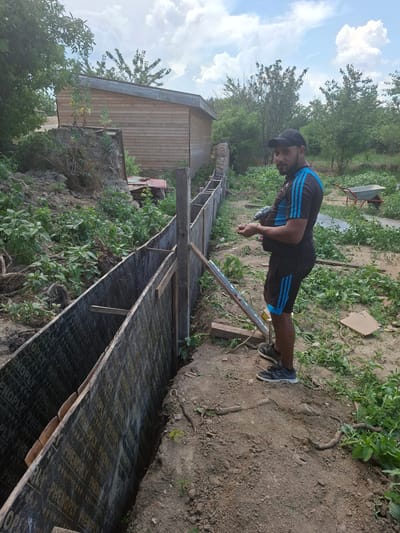 A man is working on a construction project outdoors in the city of Dolna Malina, Bulgaria. He is standing next to a wooden formwork structure that appears to be part of a foundation or retaining wall. The formwork is made of dark plywood with text that appears to be "PHENOPLY". The man is wearing a black and blue Adidas tracksuit and a black baseball cap. He is holding a drill and appears to be in the process of working on the formwork. The surrounding environment is overgrown with vegetation, and a wooden building is visible in the background. A wheelbarrow is also present in the distance. The sky is partly cloudy.