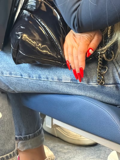 The media file shows a close-up, overhead view of a person sitting, focusing on their lap and hands. The person is wearing blue denim jeans and a dark blue jacket. They have long, bright red acrylic nails, with one nail on their left hand adorned with a white floral design. The person is holding a shiny black bag with a chain strap, which is resting on their lap. The background consists of a blue textured surface, likely a seat, and a glimpse of a white shoe. The lighting suggests daytime. There is no visible text. The overall impression is of a person in a casual setting, possibly traveling or waiting. The scene is set in Bittsa, Russia.