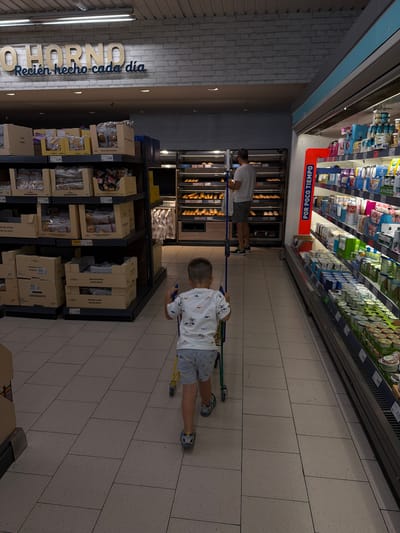 This image, taken in what appears to be a supermarket or grocery store aisle, is rotated 90 degrees counter-clockwise. The setting is an indoor commercial space with bright artificial lighting. The floor is covered with light-colored, square tiles.

In the foreground and center, a young child, likely a boy, is seen from behind, pushing a small shopping cart with a blue handle. He is wearing a white t-shirt with a pattern, light-colored shorts, and blue and white sneakers. His gaze is directed forward and slightly to the right.

Further down the aisle, towards the left, an adult man is visible, also from behind, bending over a display. He is wearing a white t-shirt and grey shorts. He appears to be stocking or organizing items in a bakery section, which features several shelves filled with bread and pastries.

Along the left side of the aisle, there are multiple shelves displaying packaged goods, including various baked items in cardboard boxes. The wall behind these shelves has a light grey brick-like texture with a prominent sign that reads "O HORNO" (partially visible, likely referring to "The Oven") and "Recién hecho cada día" ("Freshly made every day").

On the right side of the aisle, a long, multi-tiered refrigerated display unit is visible, stocked with numerous dairy products like yogurts. Above this unit, an orange sign with white text reads "POR POCO TIEMPO" ("FOR A SHORT TIME" or "Limited Time"). Various price tags are scattered throughout the shelves, indicating prices such as "1,99", "2,35", "2,69", "1,49", and "1,92". Other product labels include "MAGDALENAS" (muffins/cupcakes), "CROISSANTS", "ENSAIMADAS" (a spiral pastry), "PAN DE LECHE" (milk bread), and "BOLLOS" (buns).

The scene captures a mundane moment of shopping, with both an adult and a child navigating the store aisles. The Spanish text throughout the store confirms the location context of Spain, specifically Burjassot as provided.