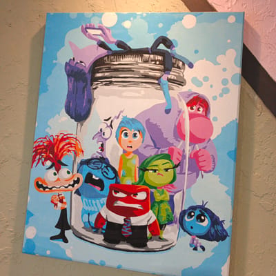 The image is a painting on canvas featuring characters from the Pixar movie "Inside Out." The characters are depicted inside a large glass jar, with a light blue, cloudy sky as the background. The characters include Joy (blue hair), Sadness (blue skin), Anger (red), Fear (purple), Disgust (green), and Bing Bong (pink elephant-like creature). Joy is in the center, looking distressed. Fear is to her left, with a terrified expression. Sadness is to Joy's right, looking concerned. Anger is in the foreground, wearing a suit and tie, with a furious expression. Disgust is to Anger's right, looking unimpressed. Bing Bong is a large figure in the background, partially obscured by the jar. The painting is displayed indoors, leaning against a textured, light brown wall. The lower right corner of the image shows a sliver of a pink wall and a dark, diagonal line, possibly part of the room's decor or another object. The painting is lit from above, casting a slight shadow. The location in Vyshgorodok, Russia is not visible in the image and cannot be determined.