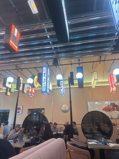 The image displays the interior of a Japanese-themed restaurant located in Clamart, France. The ceiling features an exposed industrial aesthetic with black metal tracks, from which numerous illuminated signs displaying Japanese script in various shapes (rectangular, lantern-like, circular) and colors (red, blue, yellow, orange) are suspended. White globe lights and a linear fluorescent fixture with an amber glow are also integrated into the ceiling.
The walls are clad in light wood paneling, adorned with Japanese artwork, including a circular piece depicting a red sun motif and a large framed print of a traditional Japanese figure holding a red fan. Dark-framed arched reflective panels are embedded in the walls, mirroring portions of the dining area and patrons.
The restaurant is furnished with tables featuring light-colored tops and dark edges, accompanied by diverse seating, including plush pink upholstered chairs with gold-toned frames and matching booth seating in the foreground. Several individuals are seated at tables. In the mid-ground, a woman wearing a striped shirt and a light brown hijab is actively viewing content on a smartphone held with both hands. She is seated across from another woman with long dark hair wearing a black top. Their table holds multiple drinks, including yellow liquids in glasses, and a blue ribbed plastic bottle. Further within the restaurant, a bald man in a dark shirt is seen in profile, raising a glass to his lips, while other patrons are visible dining and conversing. The scene conveys a casual, modern dining environment with pronounced East Asian decorative elements.