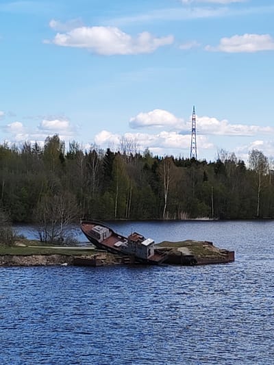 The image shows an old, possibly wrecked, vessel resting on a small, grassy island in a body of water. The vessel appears to be partially submerged or tilted significantly. The surrounding water is choppy with a blue hue. In the background, a forested area is visible with trees of varying green shades. A tall, thin, blue-and-red lattice tower rises above the treeline. The sky is blue with scattered white clouds. The location is Ivanov Bor, Russia.