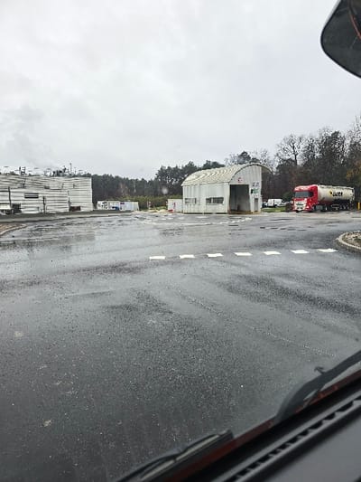 The image depicts an industrial or commercial yard in Haguenau, France, under an overcast sky, with the ground wet from recent precipitation. The perspective is from inside a vehicle, evidenced by the dashboard and windshield wipers in the lower frame. The foreground shows dark, wet asphalt with white dashed lines marking a road or turning area.
In the midground, a white corrugated metal building with a curved roof and an open archway entrance stands prominently. To its immediate right, a red semi-truck with a white tanker trailer is parked, oriented towards the right side of the frame. The tanker bears dark lettering that appears to read "JIKE" or "JAKE." To the left of the curved building, a large stack of white, rectangular structures or materials is visible. Further in the background, additional smaller white structures and containers are present.
The background is dominated by a dense line of trees, indicating a wooded area or forest bordering the facility. No individuals are visible within the scene.