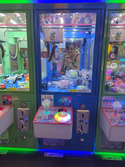This is a medium shot of a woman reflected in the glass of three connected arcade claw machines. The woman, wearing glasses and a dark tank top, is holding a mobile phone, likely taking a selfie. The claw machines are brightly lit with various colorful toys inside. The machine on the left is green and filled with toy cars. The central machine is blue and contains smaller plush toys. The machine on the right is also green and appears to contain plush toys and other prizes. The overall environment appears to be an indoor arcade. The image is a photograph taken from a low angle, looking up at the machines and the reflection.