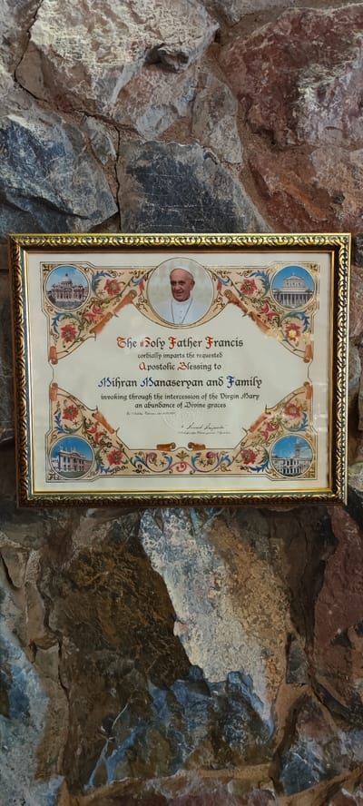 The framed certificate, adorned with ornate gold trim, is centrally positioned against a rough, natural stone wall. The certificate displays a portrait of Pope Francis at the top, with decorative architectural vignettes on either side. The text within the certificate reads: "The Holy Father Francis cordially imparts the requested Apostolic Blessing to Mihran Hanaseryan and Family invoking through the intercession of the Virgin Mary an abundance of Divine graces." Below this, there is handwritten text, including a signature and a date of "18.02.2017". The certificate is illuminated by natural light, casting subtle shadows on the textured stone background. The overall scene suggests a place of religious or historical significance, possibly a church or monastery, given the subject matter and the evident antiquity of the stonework. The location is identified as Taperakan, Armenia.