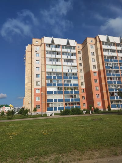 The image shows a tall, multi-story apartment building with a facade of light beige brick and orange-pink sections. The building has many balconies with blue and white railings. The sky above is a vibrant blue with scattered white clouds. In the foreground, there is a grassy area with some small yellow wildflowers. To the left, in the distance, other apartment buildings and a green-roofed structure are visible. A single person wearing a reddish-orange jacket and dark pants is walking on a pathway in front of the building. The scene appears to be in a residential area of Polatsk, Belarus.